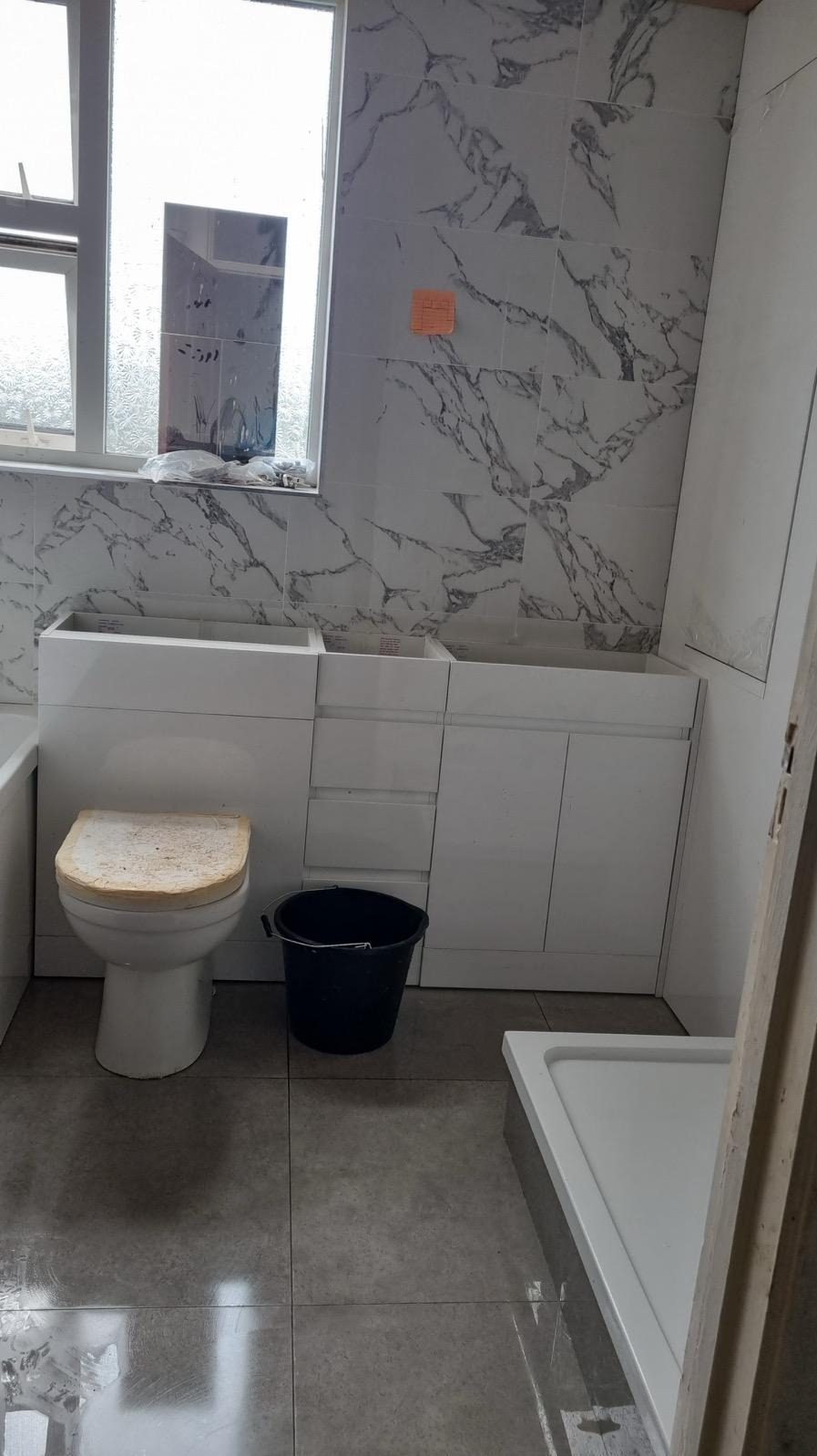 Łazienka w trakcie prac: białe marmurowe płytki, zabudowa umywalki z szafkami, toaleta z zabrudzoną deską, szare płytki podłogowe i czarne wiadro. Widok na okno.
