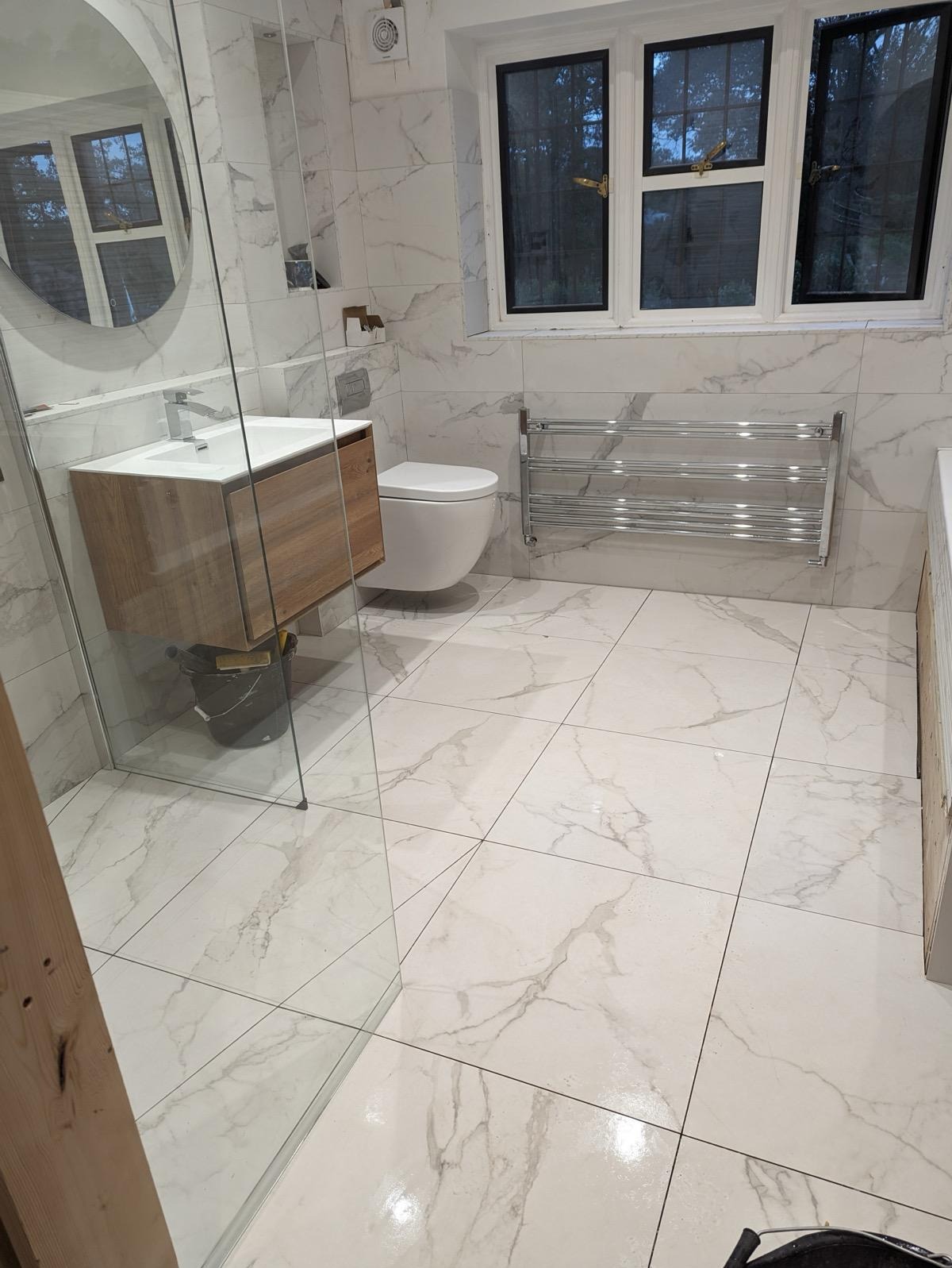 Wykończona łazienka z marmurowymi płytkami, wiszącą szafką z umywalką, toaletą i grzejnikiem drabinkowym. Kabina prysznicowa ze szklanymi drzwiami. Nowoczesny design.