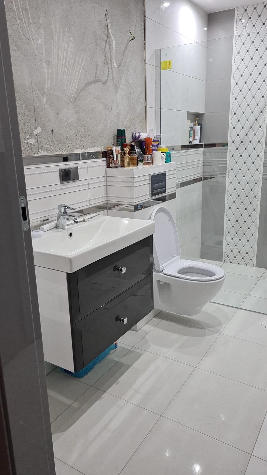 Łazienka w trakcie wykańczania: białe płytki, umywalka z szafką w odcieniach szarości, toaleta. Ściana częściowo surowa, widoczne instalacje.