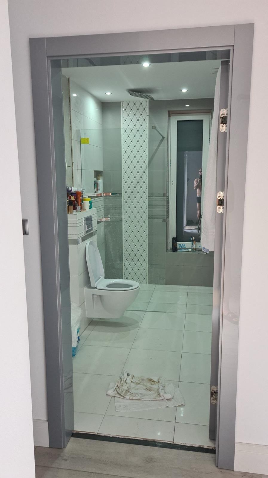 Nowoczesna łazienka z toaletą typu Geberit, widoczna przez szarą futrynę drzwiową. Białe płytki podłogowe i dekoracyjne panele ścienne. W tle okno z odbiciem osoby.