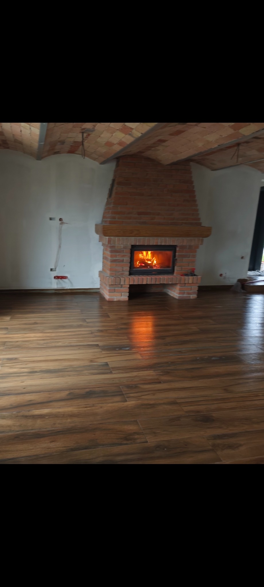 Ukończony salon z drewnopodobną podłogą i ceglanym kominkiem z paleniskiem. Widoczne niedokończone ściany i sufit z ceglanymi łukami.