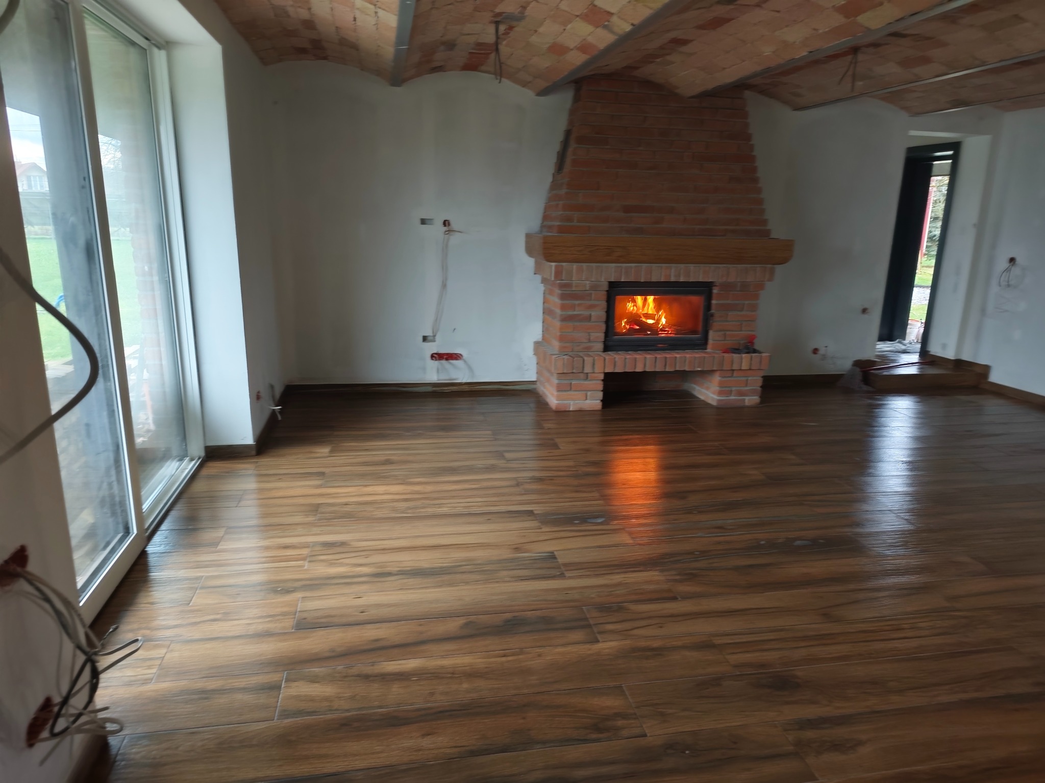 Salon z ceglanym kominkiem i drewnopodobną podłogą. Widoczne duże okno i sufit z cegieł. Ściany w trakcie wykończenia, widoczne kable. Ogień w kominku.
