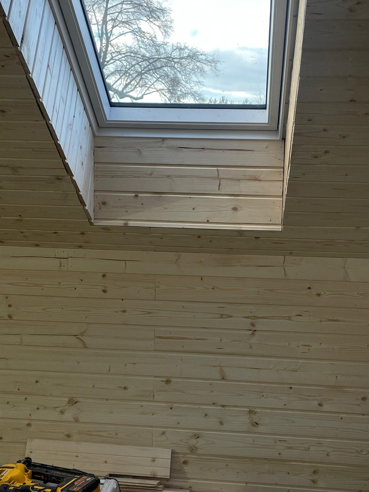 Wykończenie poddasza boazerią z jasnego drewna wokół okna dachowego. Widoczne narzędzia pracy w dolnym rogu. Naturalne światło dzienne.