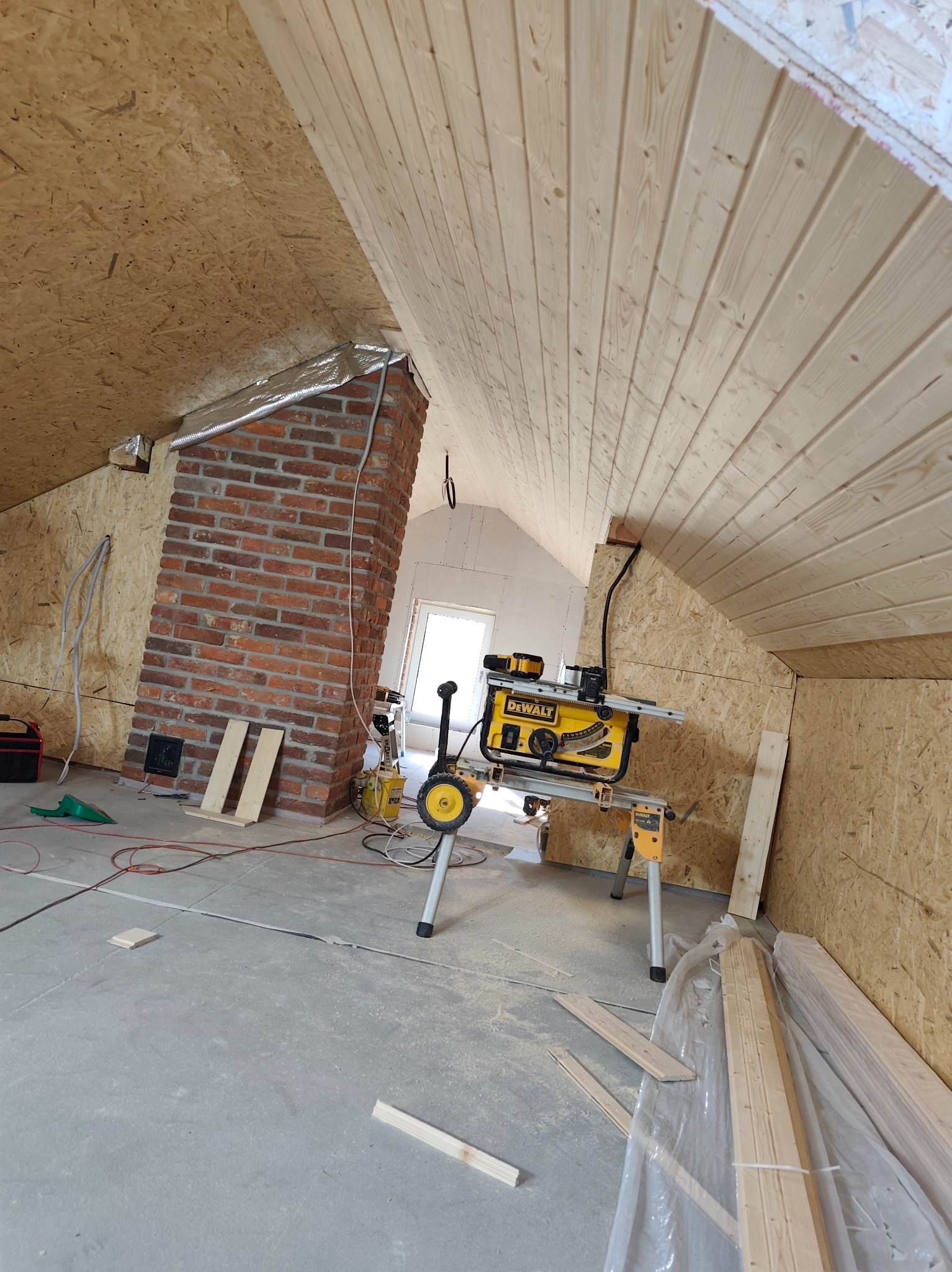 Poddasze w trakcie prac wykończeniowych: boazeria na skosach sufitu, ceglany komin, narzędzia do obróbki drewna. Widoczne płyty OSB na ścianach.