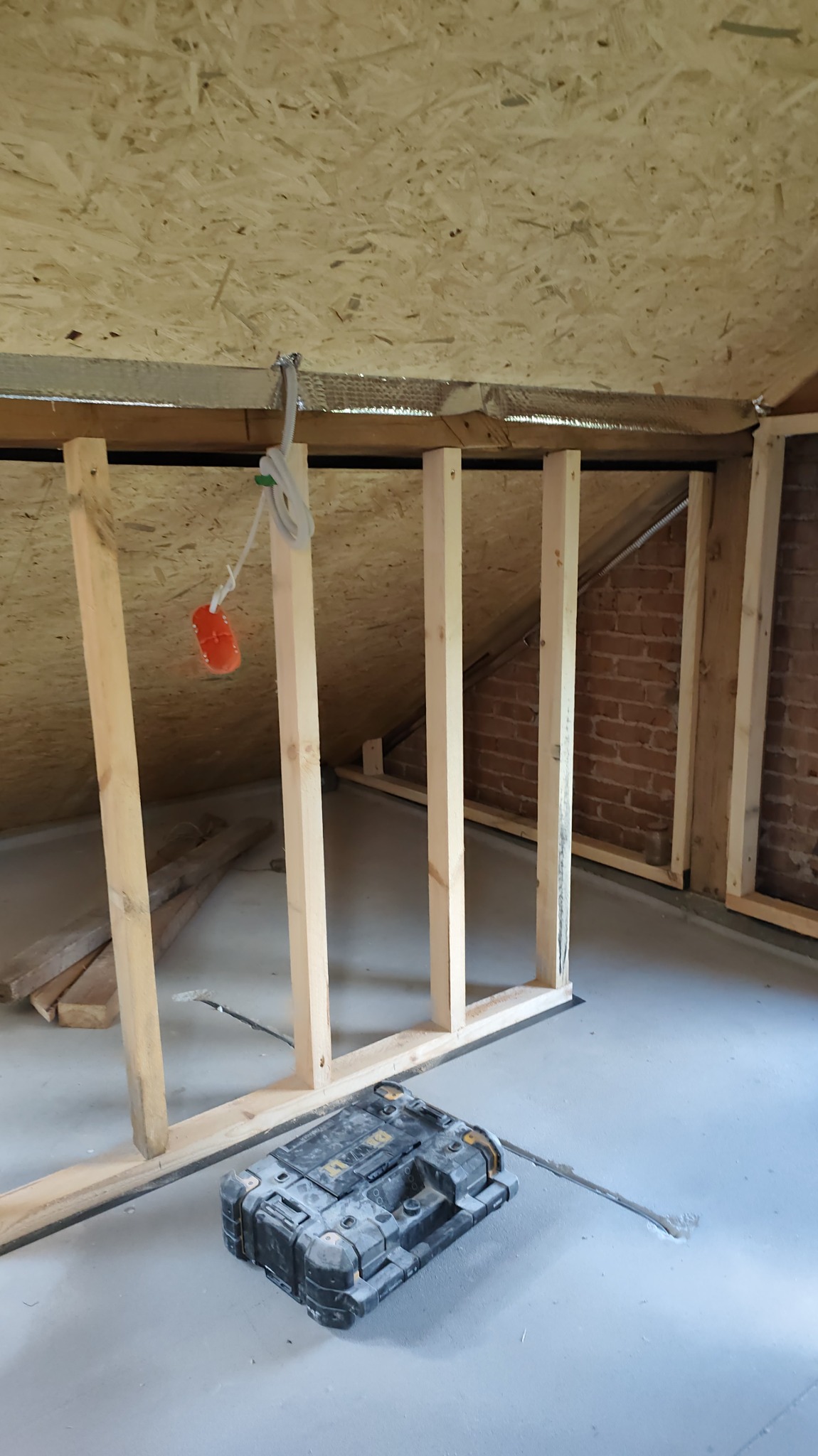 Szkielet ścianki działowej z drewnianych belek w surowym wnętrzu poddasza, widoczna ceglana ściana i skrzynka narzędziowa na podłodze.