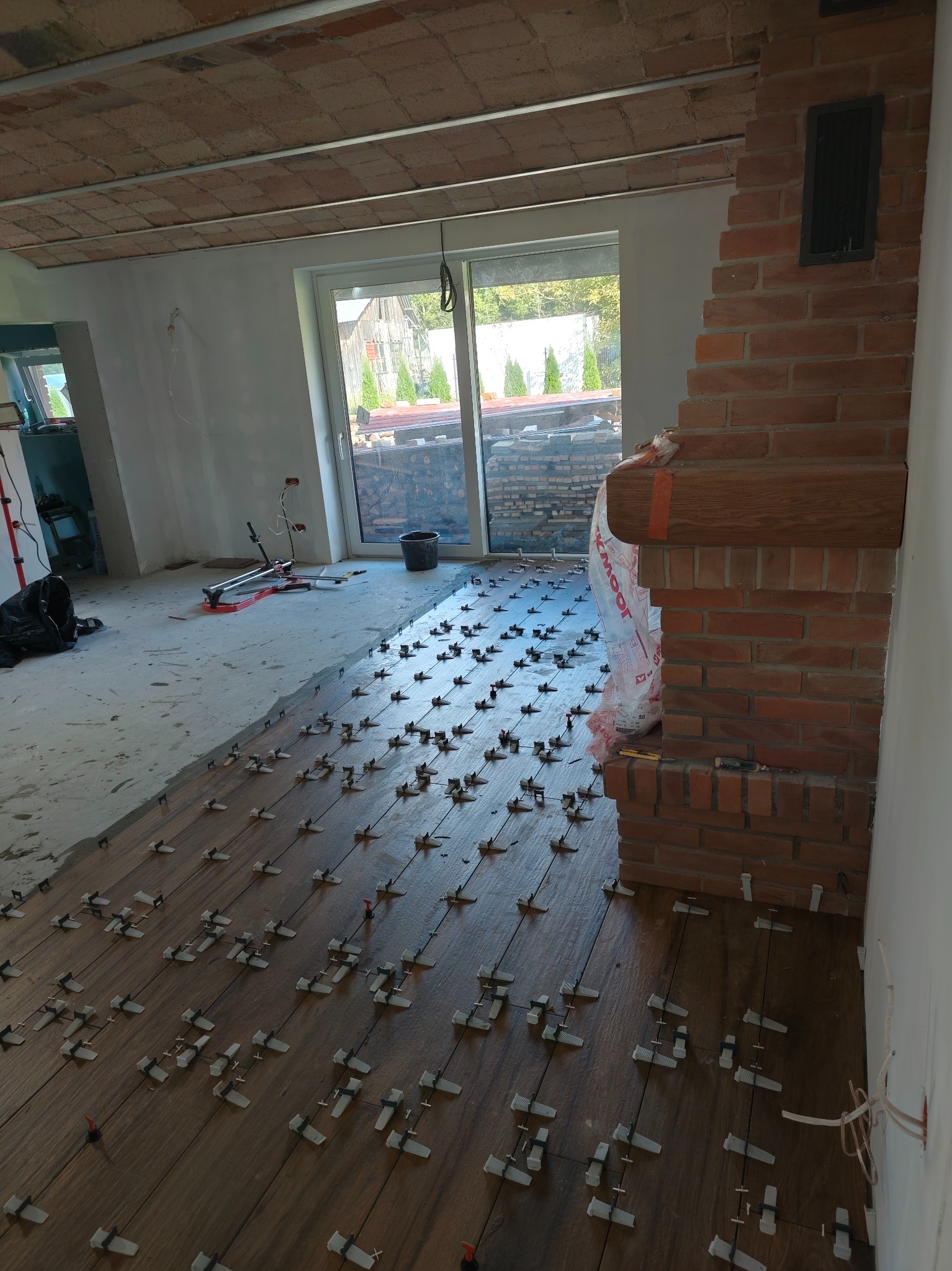 Wnętrze w trakcie prac: brązowe płytki podłogowe ułożone z użyciem klipsów poziomujących, ceglany kominek, surowy sufit i ściany w trakcie przygotowania.
