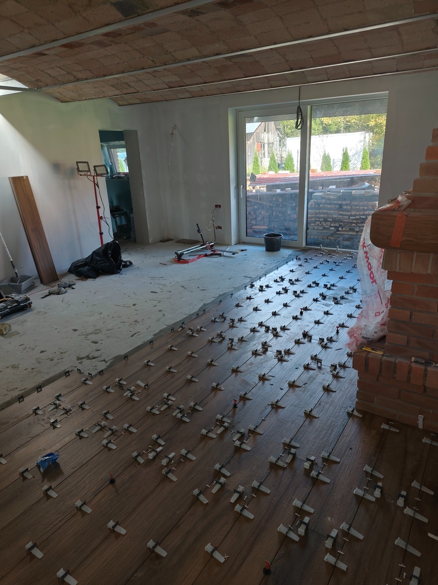 Wnętrze w trakcie prac: brązowe płytki podłogowe z systemem poziomowania, widoczny fragment kominka z cegły, okno z widokiem na zewnątrz i przygotowane materiały budowlane.