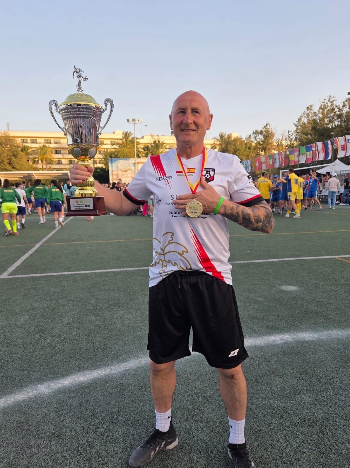 Uśmiechnięty mężczyzna w stroju sportowym trzyma puchar i złoty medal na tle boiska z flagami, świętując zwycięstwo w zawodach sportowych.