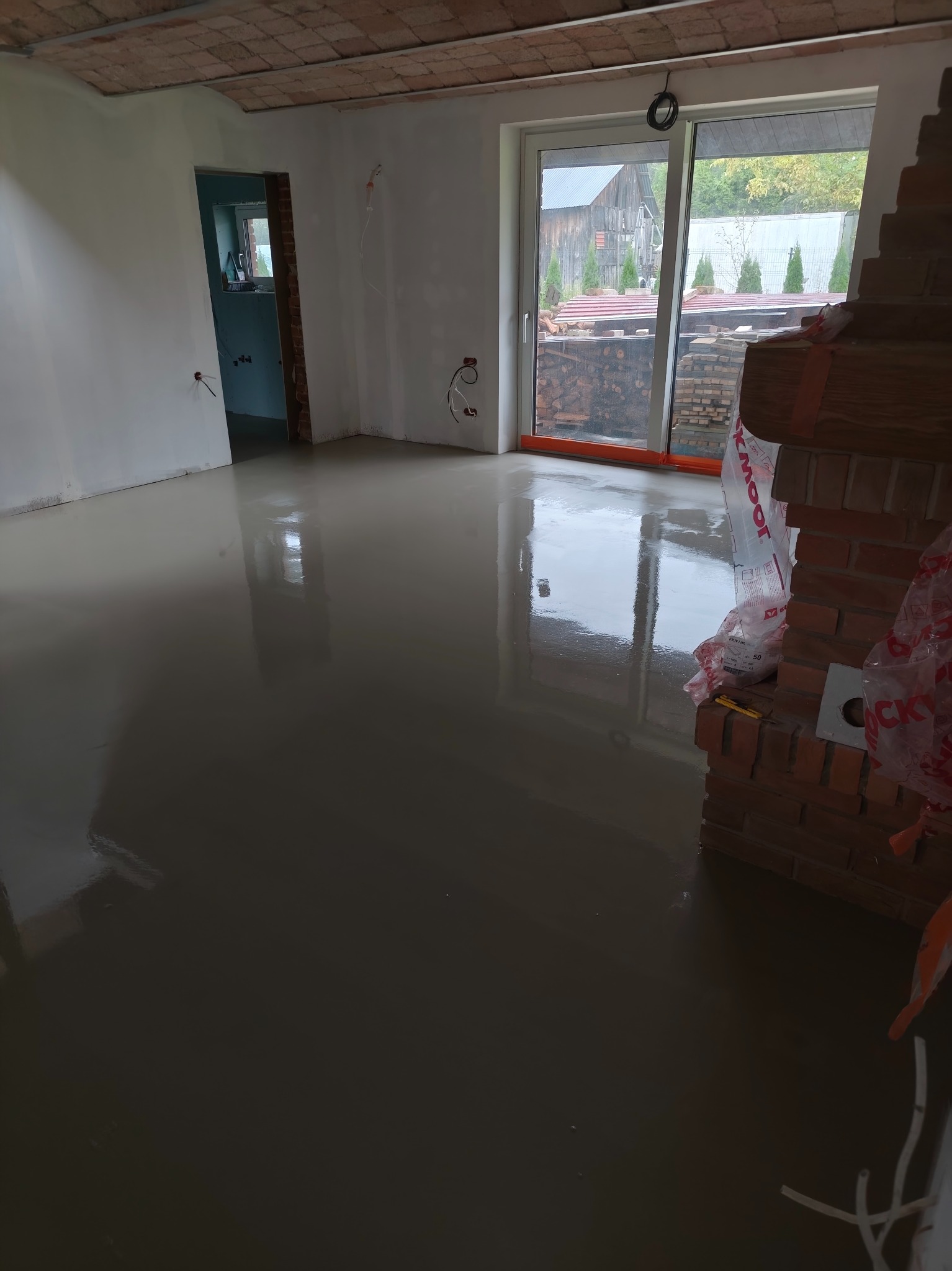 Wylany, mokry samopoziomujący podkład podłogowy w jasnym pomieszczeniu z ceglanym kominkiem i widokiem na zewnątrz przez duże okno przesuwne.