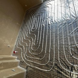 HydroPro Jakub Hereda - Białe rury ogrzewania podłogowego ułożone na czarnej siatce, widok z góry, jasne światło z okna. Etap instalacji przed wylewką.