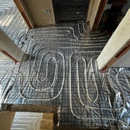 HydroPro Jakub Hereda - Białe rury ogrzewania podłogowego ułożone na czarnej siatce, rozłożone na srebrnej folii izolacyjnej w trakcie instalacji. Widok z góry na kilka pomieszczeń.