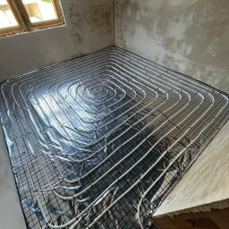 HydroPro Jakub Hereda - Białe rury ogrzewania podłogowego ułożone spiralnie na srebrnej folii izolacyjnej i czarnej siatce w surowym wnętrzu z oknem dachowym.