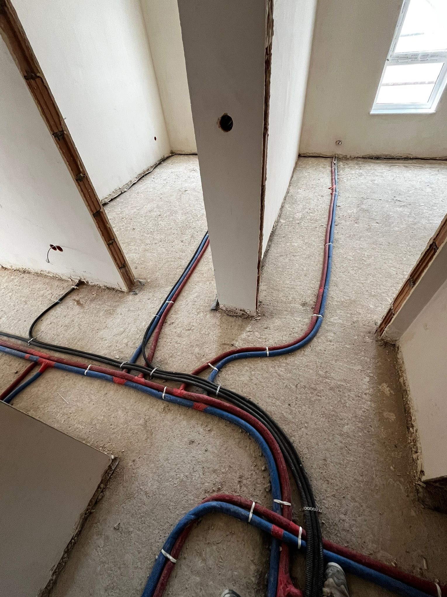 Rozprowadzone rury instalacji wodnej (czerwone i niebieskie) oraz peszle elektryczne na podłodze w surowym stanie, przed wylaniem posadzki. Widok z góry.