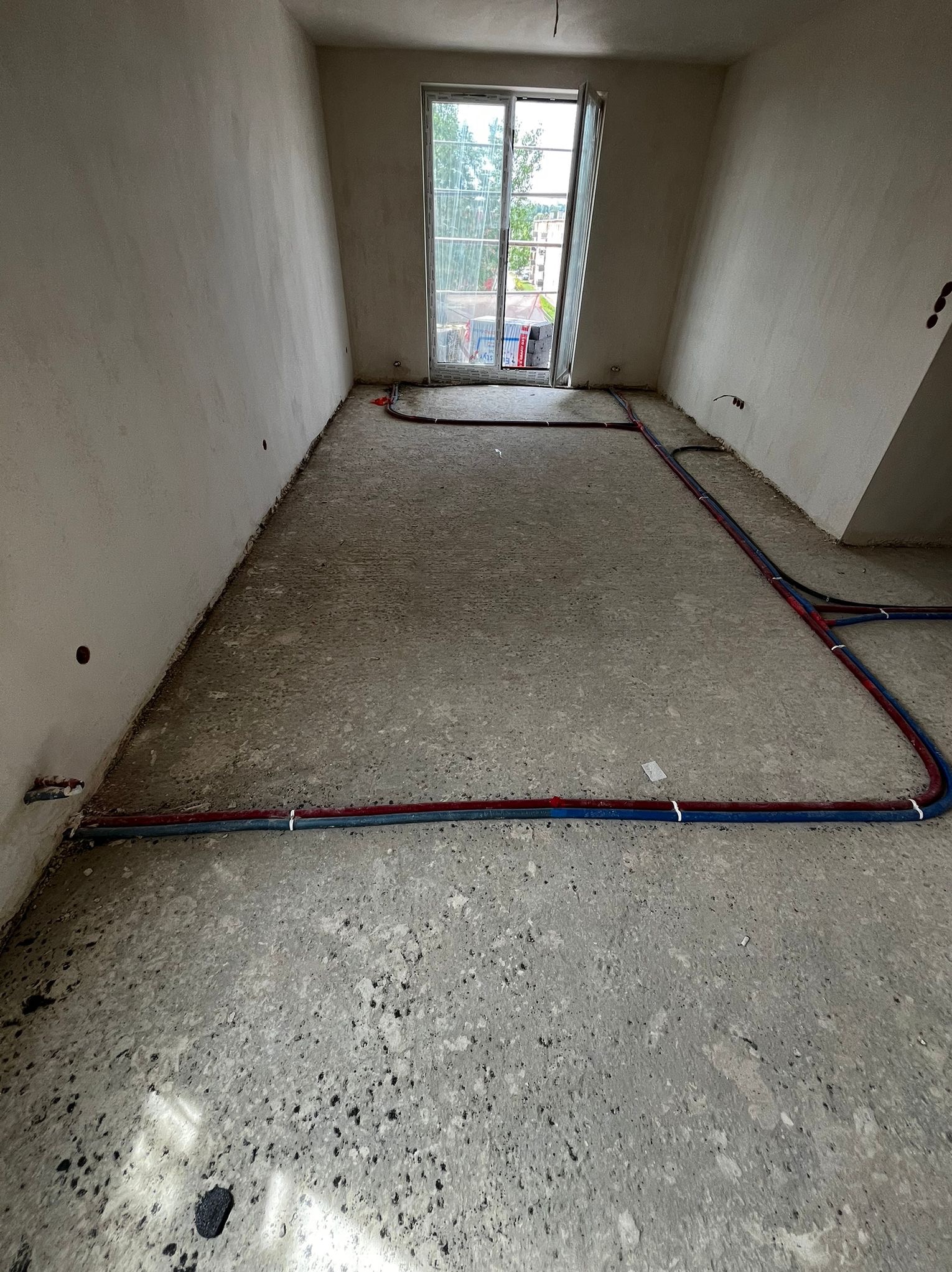 Surowy stan: Instalacja ogrzewania podłogowego z rurami czerwonymi i niebieskimi na szarym betonie, widok z góry, okno w tle, ściany w stanie deweloperskim.