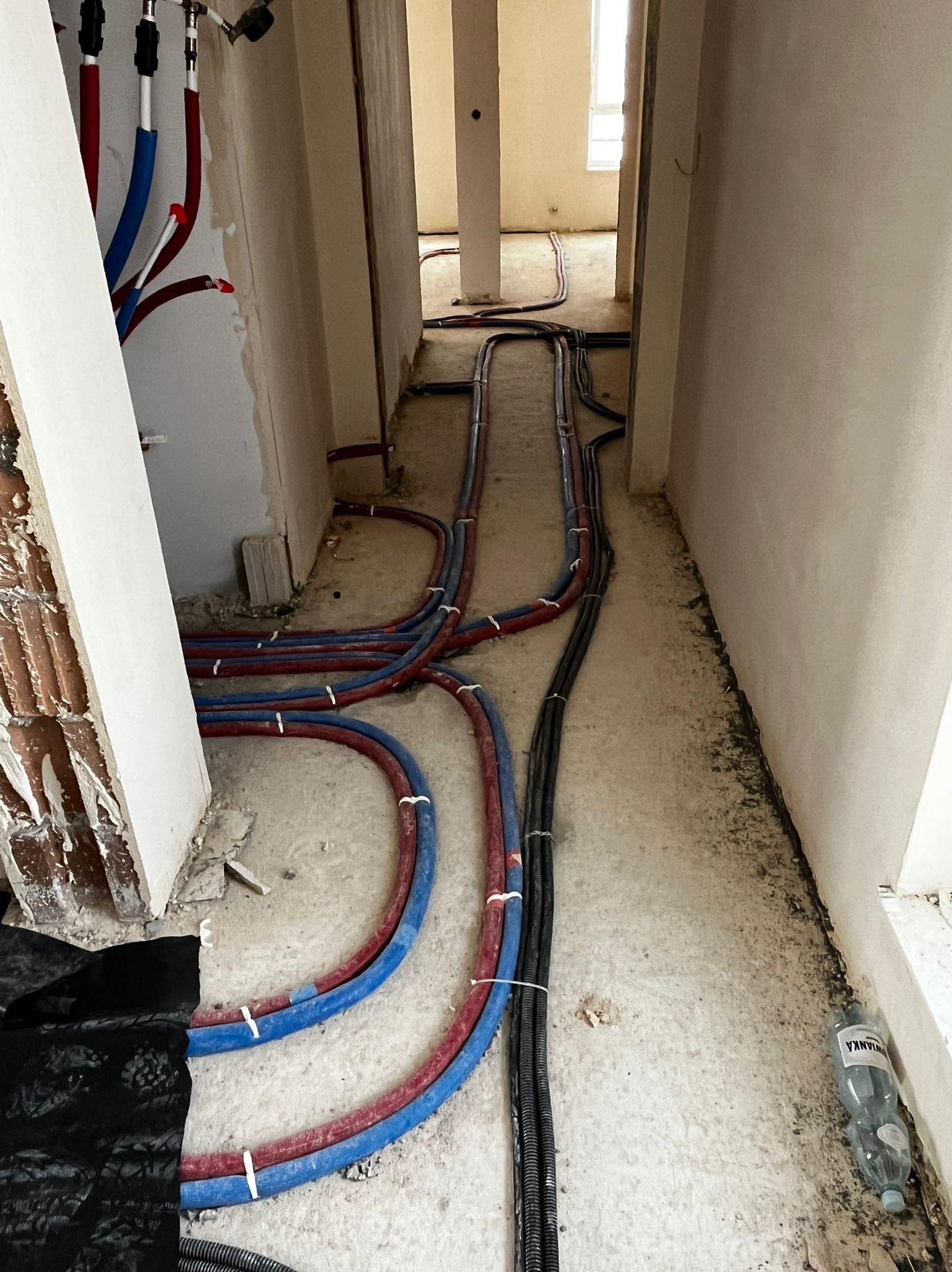 Instalacja wodna i elektryczna w trakcie montażu w surowym wnętrzu. Widoczne rury PEX w kolorach czerwonym i niebieskim oraz peszle z kablami, prowadzone po podłodze.