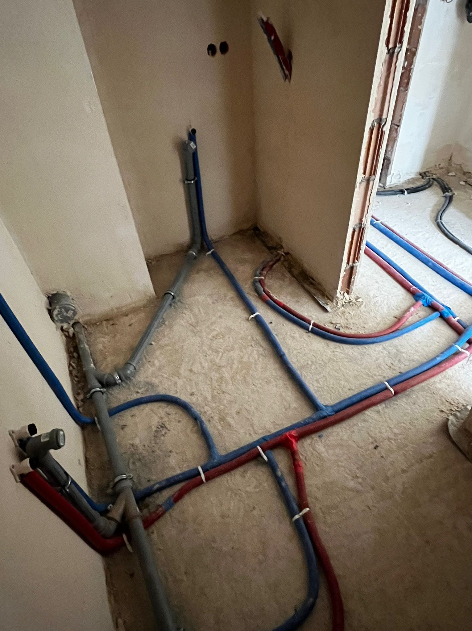 Rozłożona instalacja wodna z rurami w kolorze niebieskim i czerwonym, połączonymi białymi opaskami, na podłodze w surowym stanie. Widoczne fragmenty ścian i otwory.