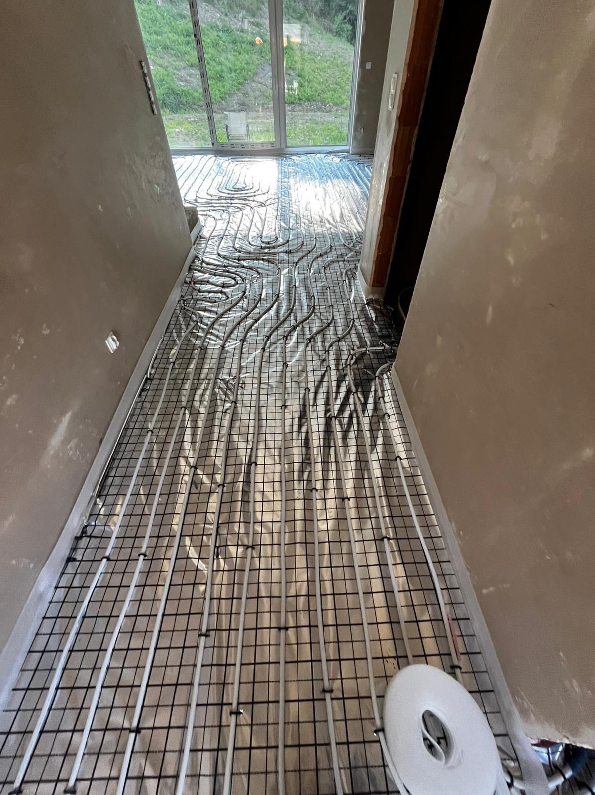 Ułożona instalacja ogrzewania podłogowego na folii, widok z góry, z rolką taśmy montażowej w rogu. W tle okno z widokiem na zieleń.