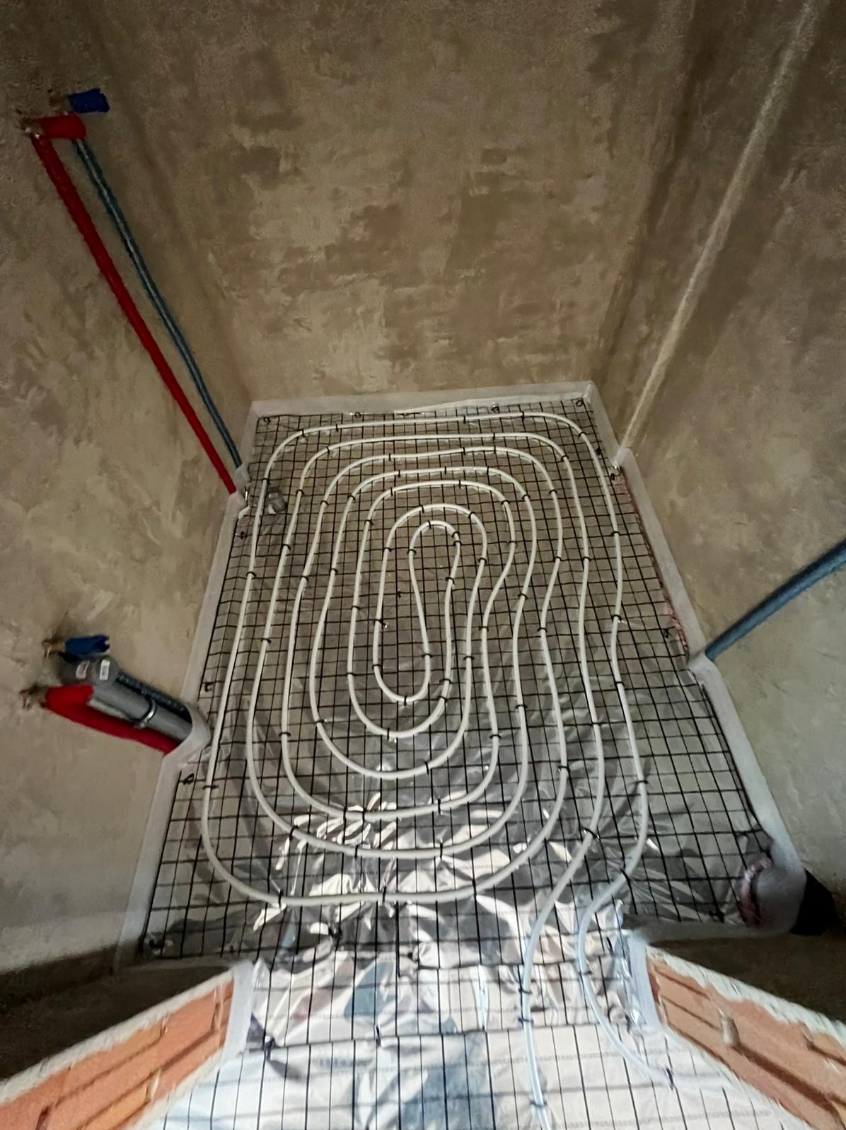 Białe rury ogrzewania podłogowego ułożone spiralnie na czarnej siatce, na folii izolacyjnej. Widoczne fragmenty ścian i rur doprowadzających wodę.