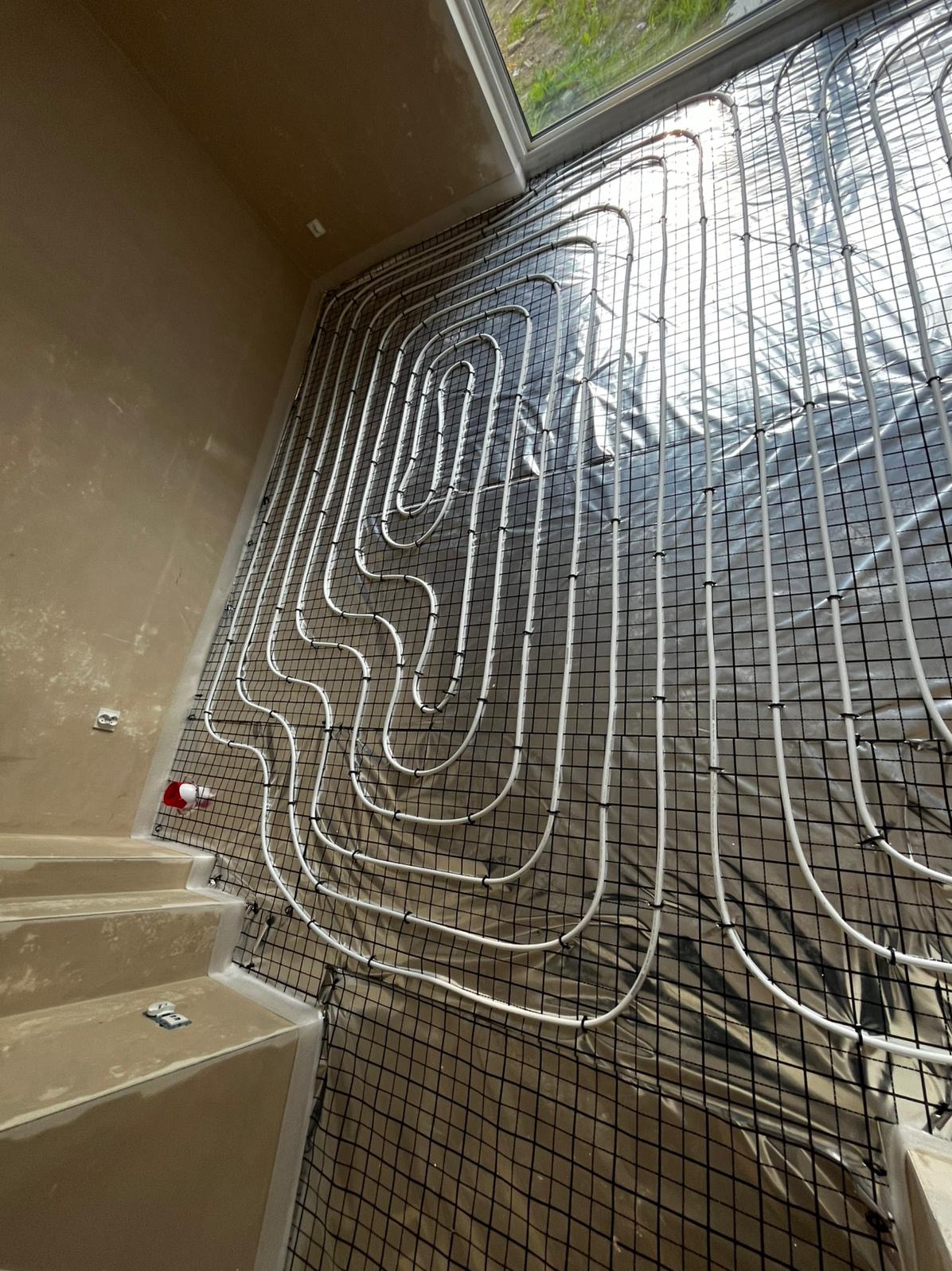 Białe rury ogrzewania podłogowego ułożone na czarnej siatce, widok z góry, jasne światło z okna. Etap instalacji przed wylewką.