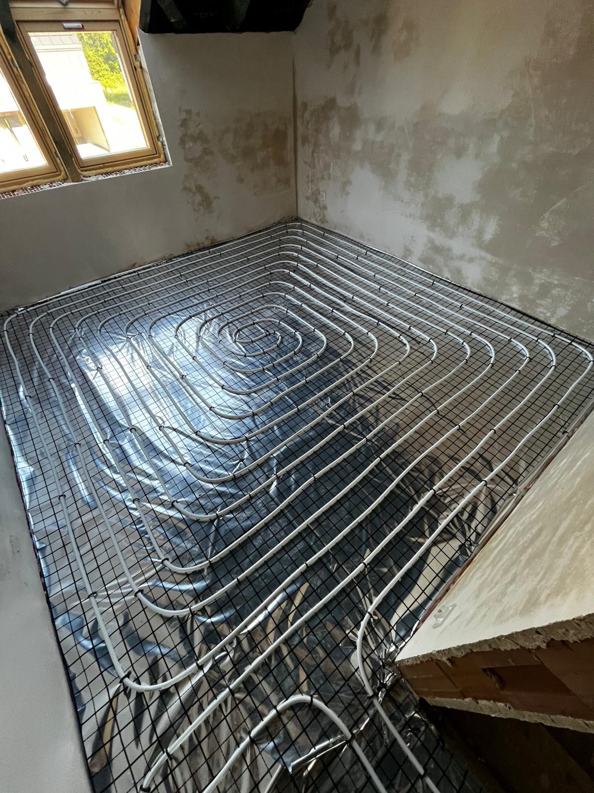 Białe rury ogrzewania podłogowego ułożone spiralnie na srebrnej folii izolacyjnej i czarnej siatce w surowym wnętrzu z oknem dachowym.