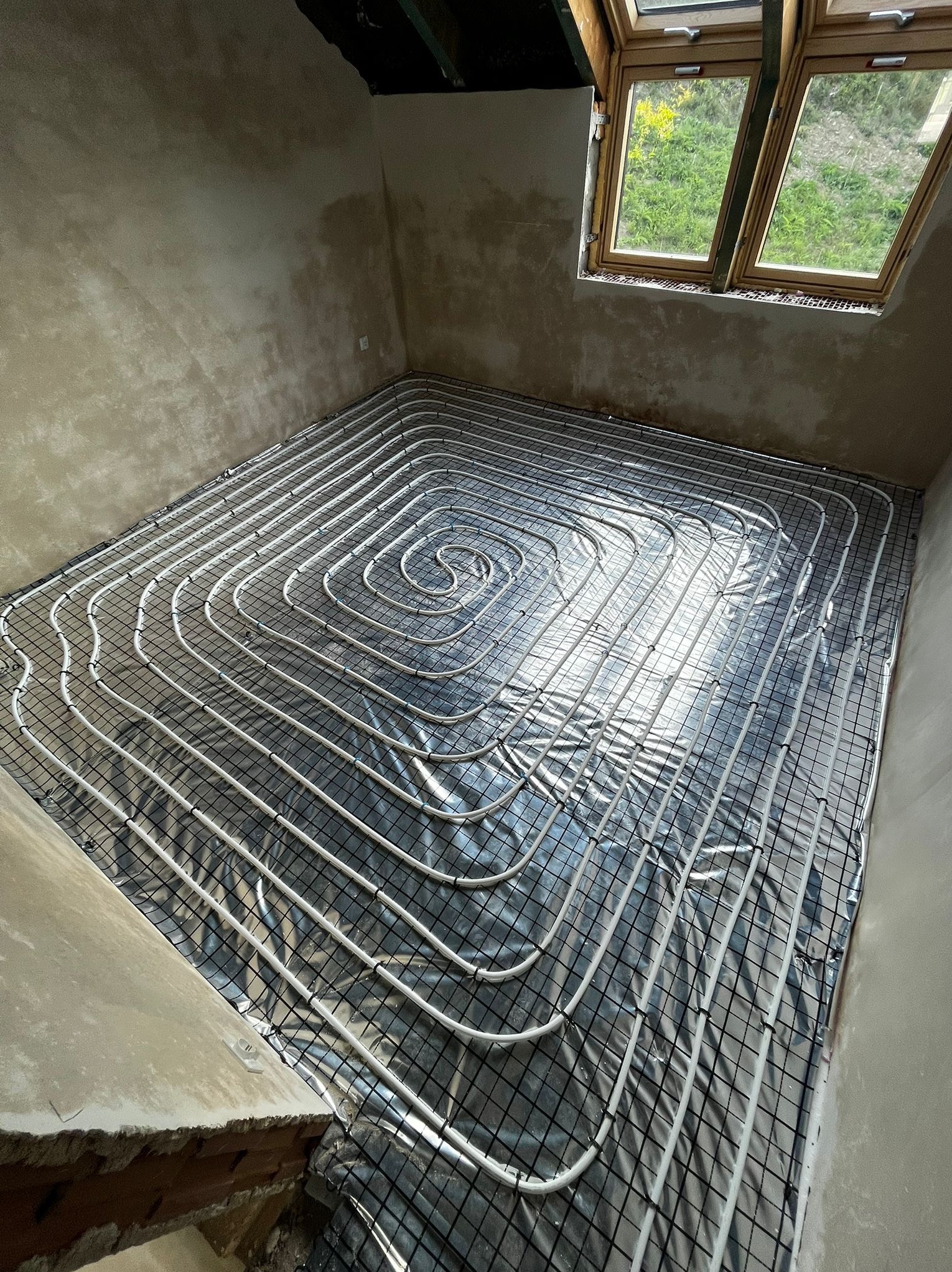 Białe rury ogrzewania podłogowego ułożone spiralnie na folii izolacyjnej i siatce w surowym wnętrzu z oknem dachowym. Widok z góry na instalację przed wylewką.