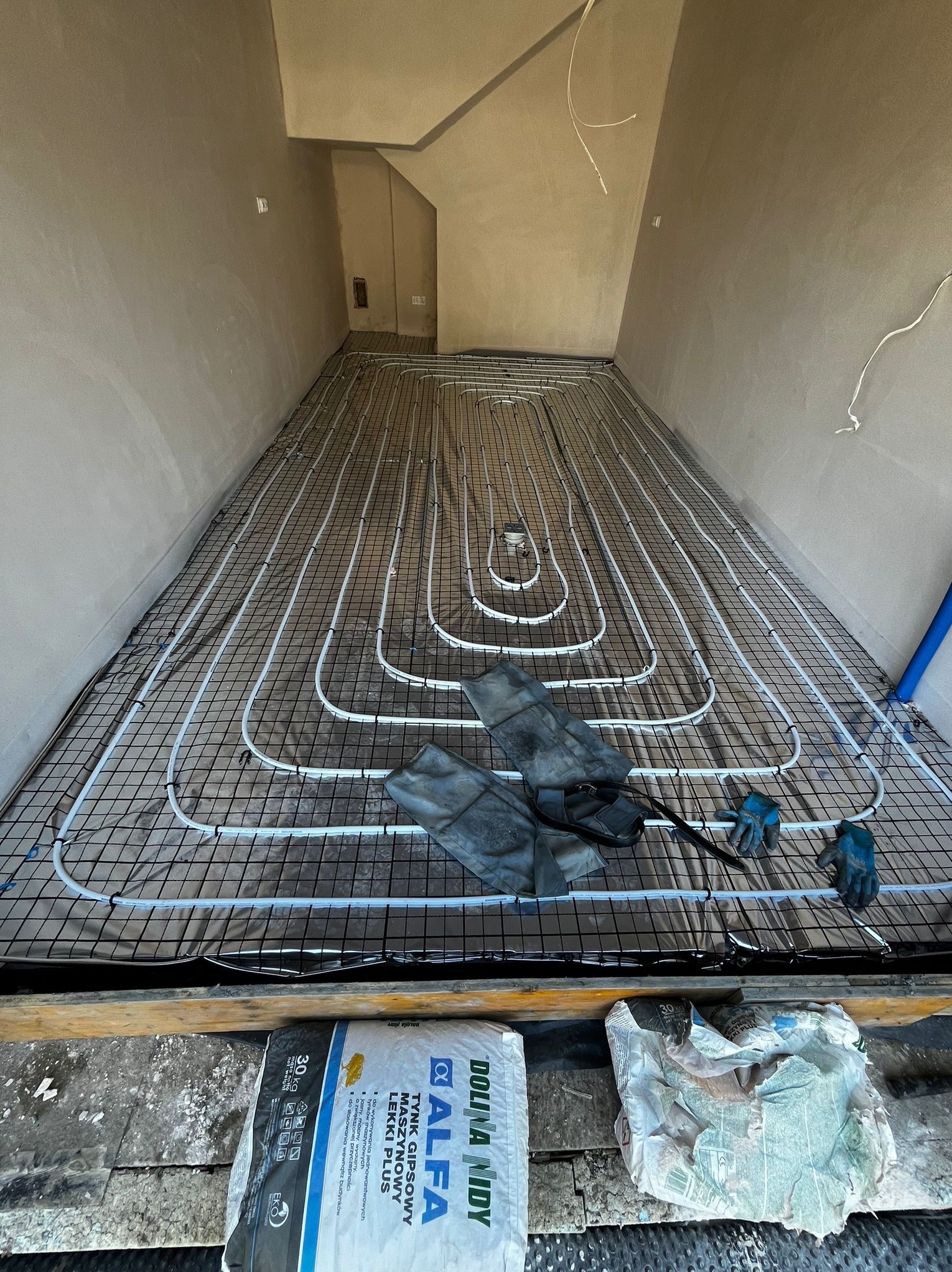 Ułożona instalacja ogrzewania podłogowego na czarnej siatce, widoczne białe rurki, rękawice robocze i worki z zaprawą. Przygotowanie do wylewki.