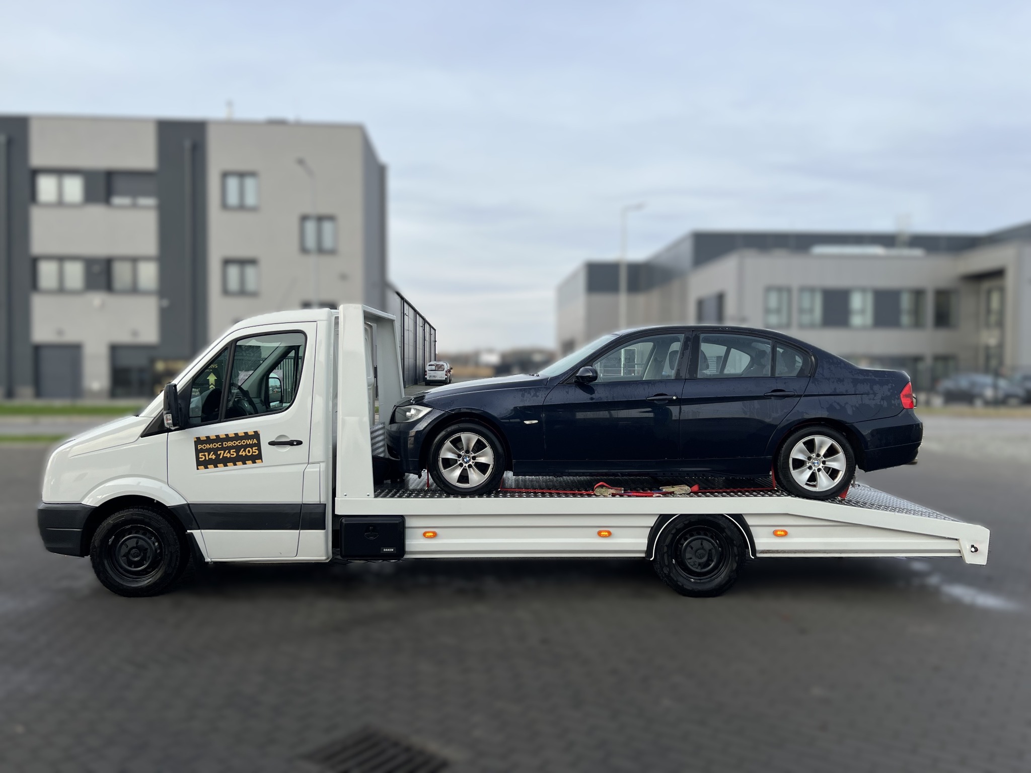 Czarny samochód osobowy na białej lawecie pomocy drogowej z numerem telefonu na tle szarego budynku. Widok z boku, pochmurny dzień.