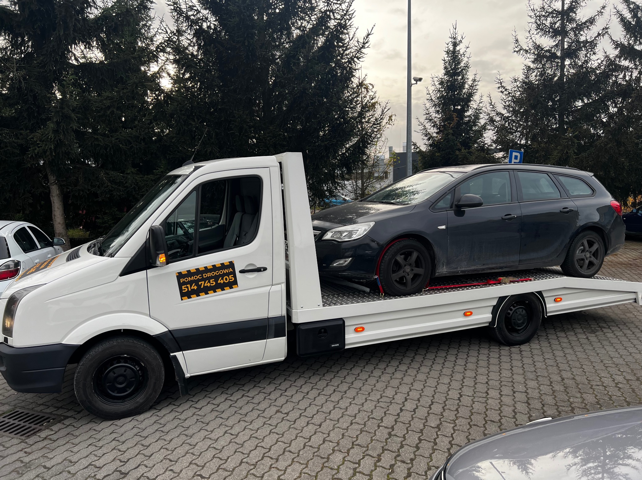 Laweta z czarnym kombi Opel Astra na platformie, zaparkowana na brukowanym placu. Na drzwiach lawety żółto-czarny napis: Pomoc Drogowa 514 745 405.