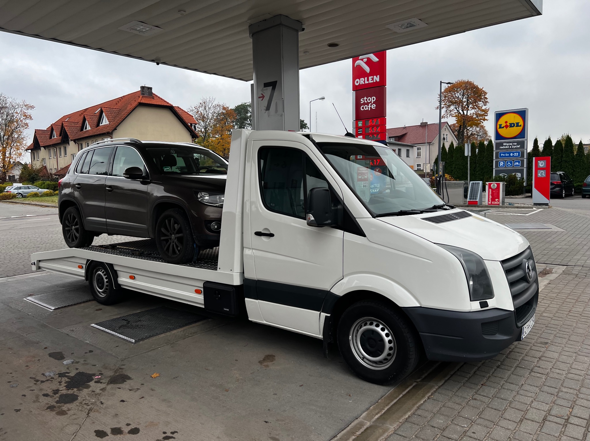 Biała laweta z brązowym SUV-em na platformie, zaparkowana na stacji benzynowej Orlen. W tle budynki i drzewa. Widoczne logo Lidl.