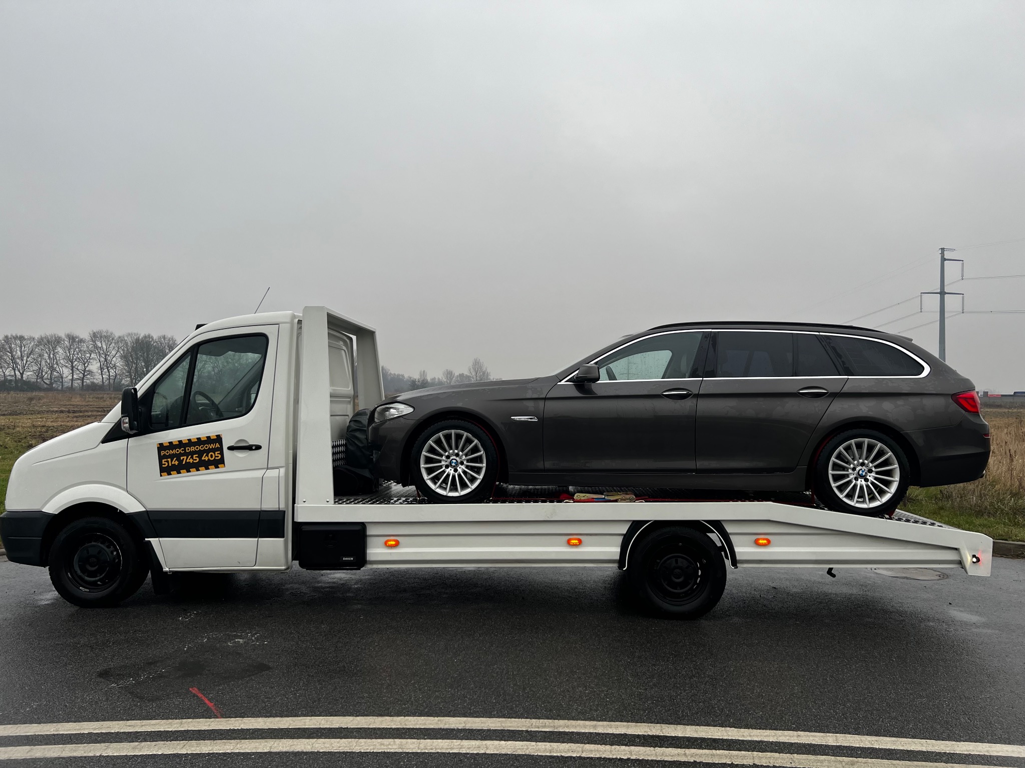 Szara BMW serii 5 kombi na białej lawecie pomocy drogowej z numerem telefonu na drzwiach, mokra nawierzchnia, pochmurne niebo.
