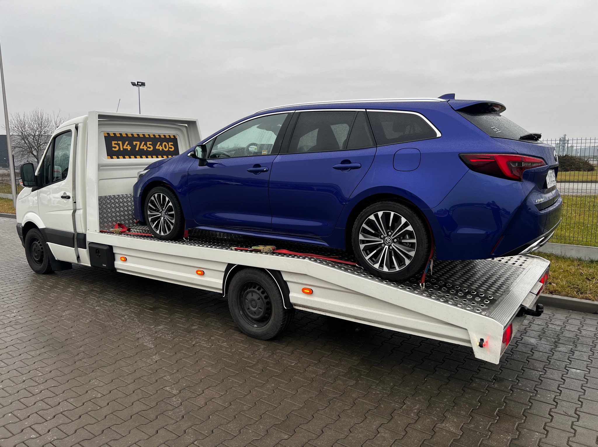 Niebieska Toyota Corolla kombi na białej lawecie z numerem telefonu 514 745 405, widok z boku na tle szarego nieba i przemysłowego krajobrazu.