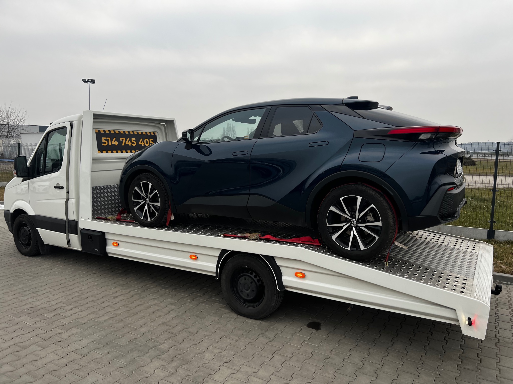 Niebieski samochód osobowy, Toyota C-HR, zabezpieczony pasami na białej lawecie z numerem telefonu, na tle szarego nieba i przemysłowego krajobrazu.