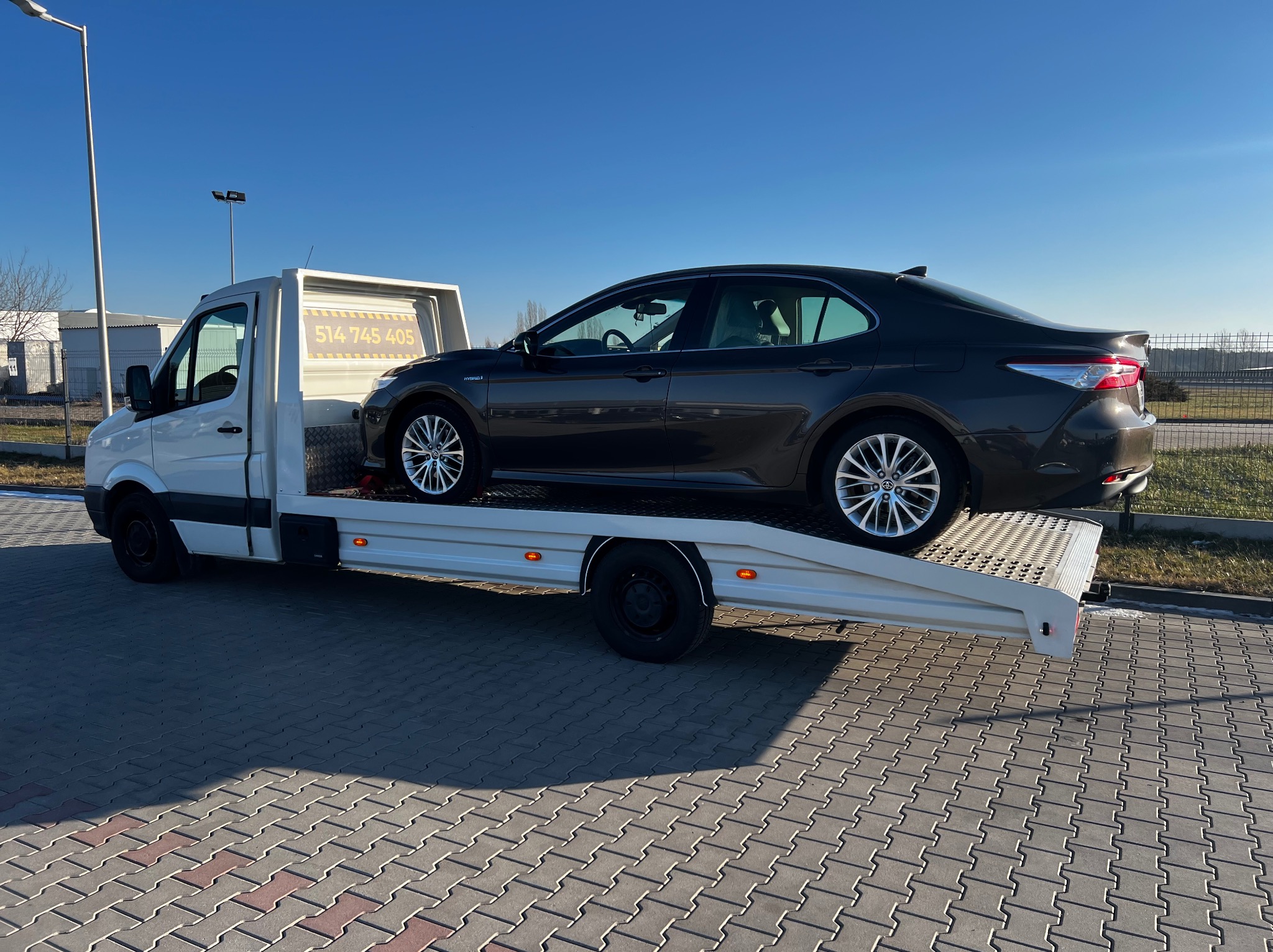 Biała laweta z czarnym samochodem osobowym na platformie, zaparkowana na kostce brukowej w słoneczny dzień. Na zabudowie lawety widoczny numer telefonu.