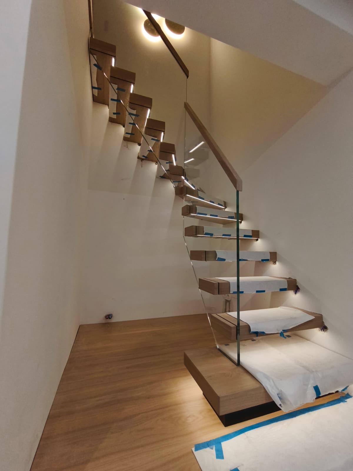 Nowoczesne schody wiszące z podświetleniem LED, zabezpieczone folią ochronną. Szklana balustrada z drewnianym wykończeniem. Minimalistyczny design, ciepłe oświetlenie.