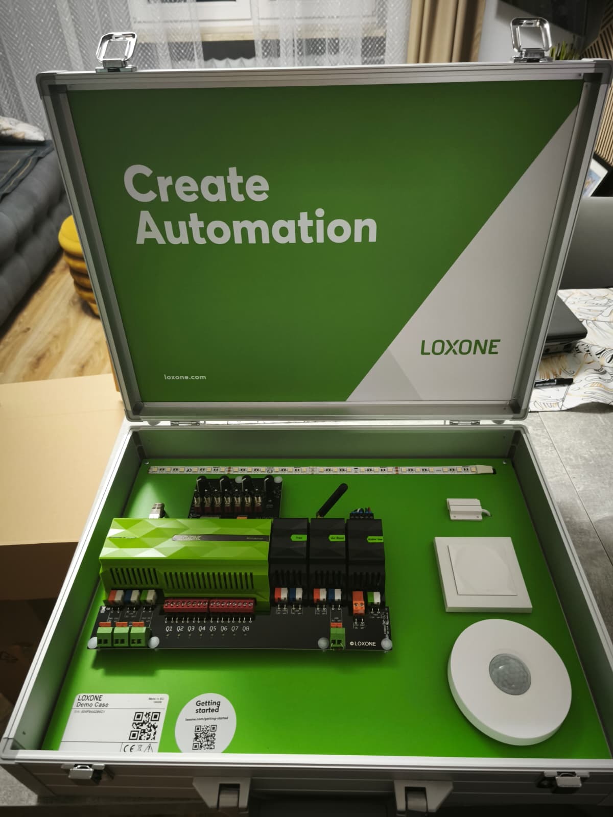 Otwarta walizka demonstracyjna Loxone z elementami automatyki domowej: centralka, moduły, czujnik ruchu i listwa LED, prezentowane na zielonym tle.