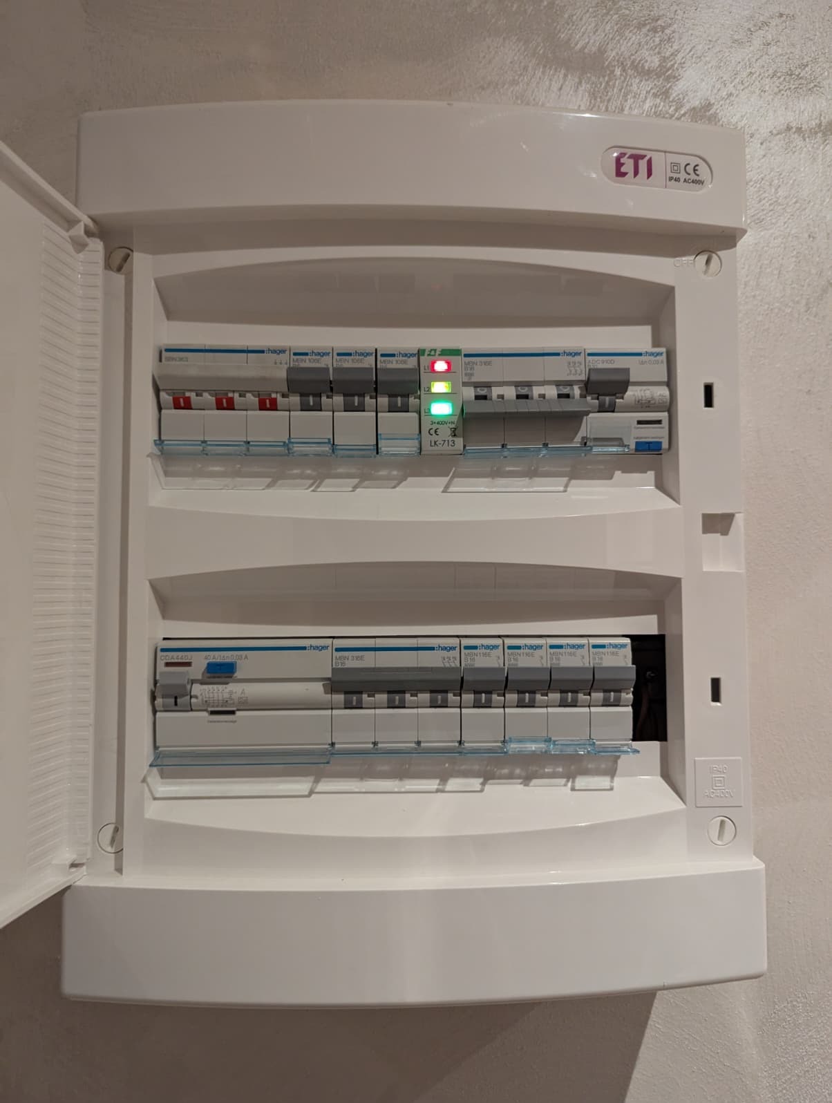 Otwarta, biała skrzynka elektryczna z bezpiecznikami Hager, widoczne diody sygnalizacyjne. Instalacja wewnątrz budynku, na ścianie. Widoczny model LK-713.