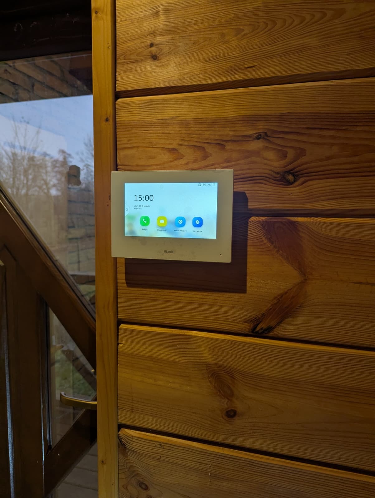 Zamontowany panel wideodomofonu Hiklook na drewnianej ścianie wewnątrz domu. Widoczny ekran z godziną 15:00 i ikonami funkcji. Obok fragment okna.