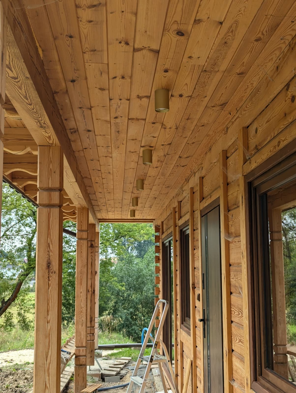 Drewniany taras z zamontowanym oświetleniem punktowym w suficie. Widoczna drabina sugeruje niedawny montaż. Naturalne światło dzienne.