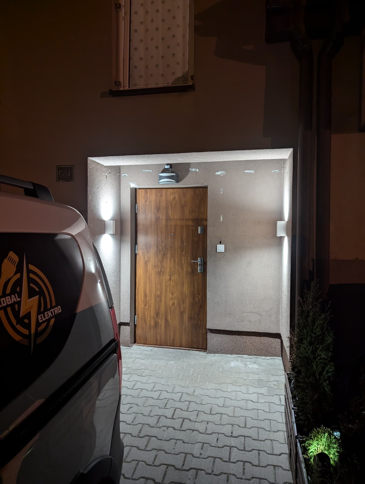 Oświetlenie elewacyjne LED przy wejściu do domu, z logo firmy elektrycznej na samochodzie w kadrze. Nowoczesne lampy ścienne po obu stronach drzwi.