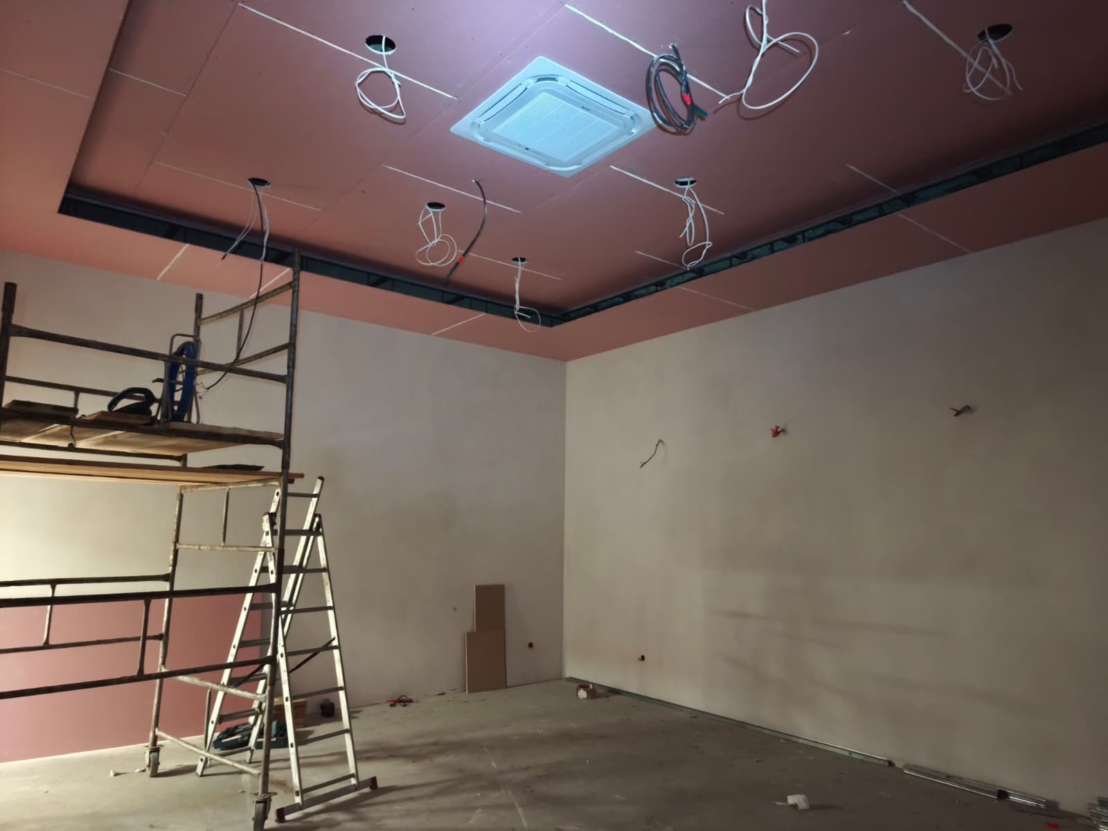 Wnętrze w trakcie prac: podwieszany sufit z klimatyzacją, wystające przewody elektryczne, rusztowanie. Widoczne przygotowania do instalacji oświetlenia.