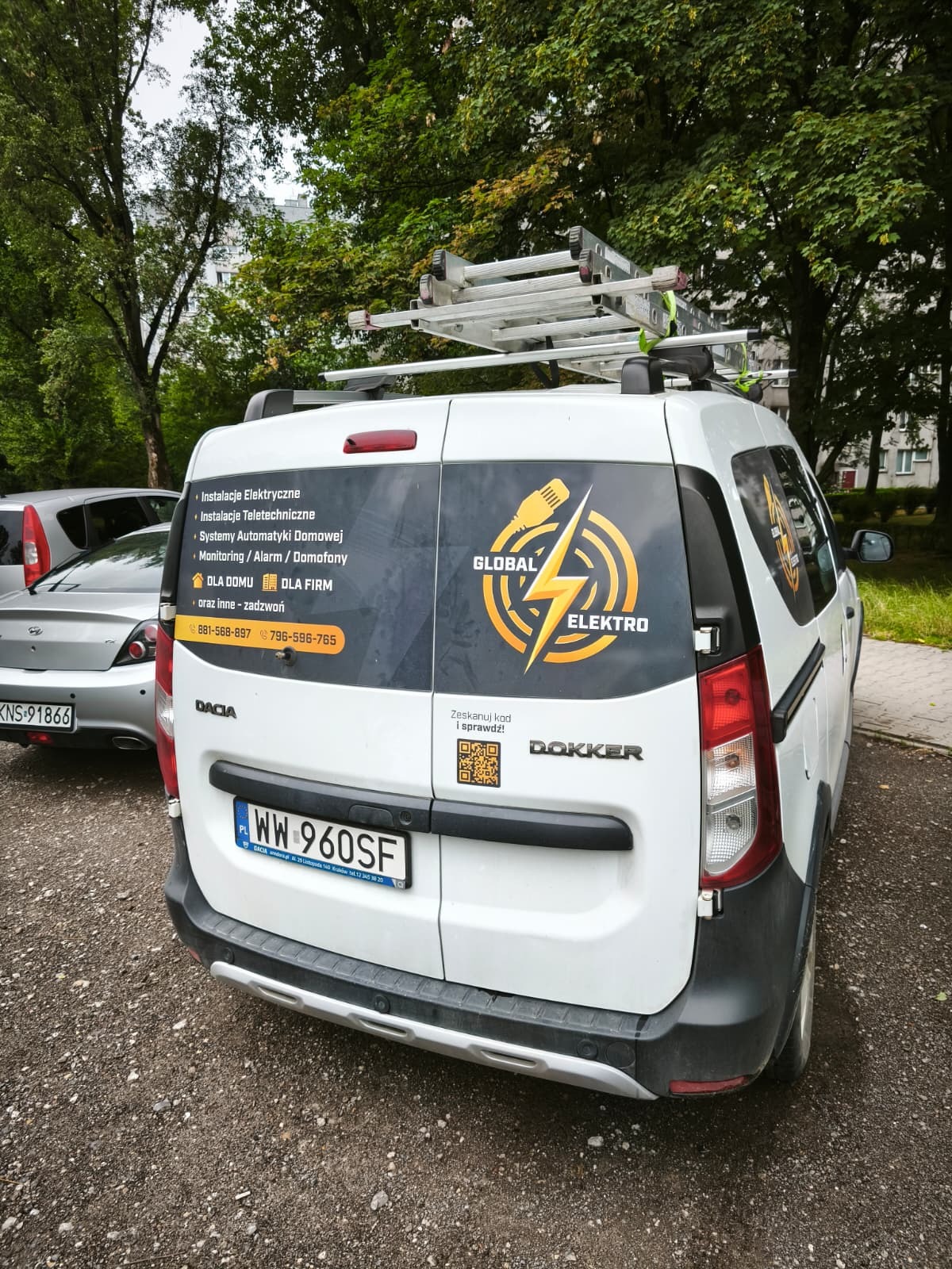 Biały samochód Dacia Dokker z logo firmy Global Elektro i listą usług: instalacje elektryczne, alarmy, domofony. Na dachu drabina. Tablica rejestracyjna WW 960SF.