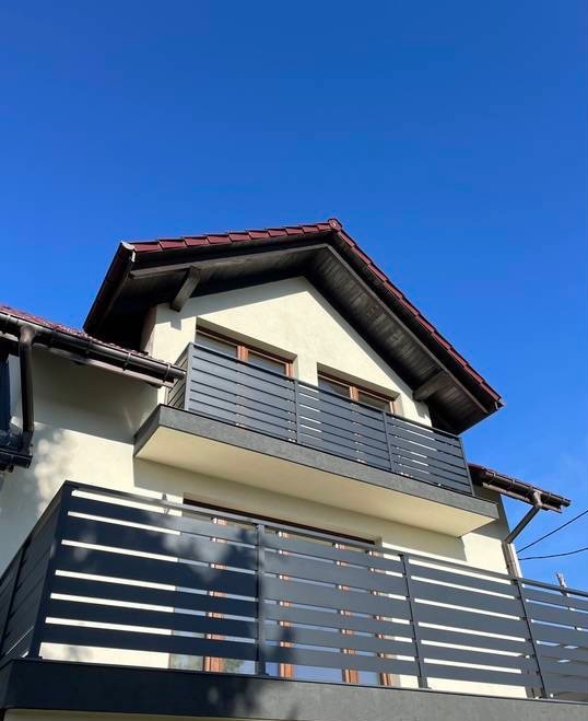 Nowoczesna balustrada balkonowa z poziomych, szarych profili na tle jasnej elewacji domu i błękitnego nieba. Minimalistyczny design, proste linie, widok z dołu.