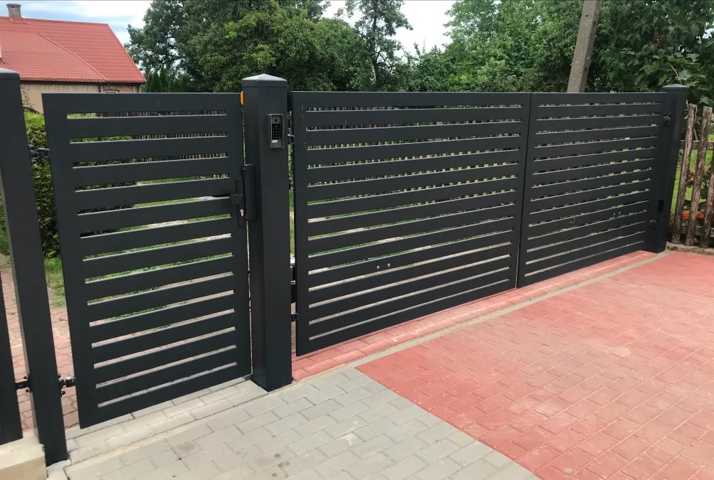 Nowoczesna, pozioma brama metalowa w kolorze grafitowym z panelem dostępowym, zamontowana na posesji z kostką brukową w odcieniach szarości i czerwieni.