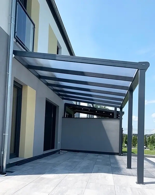 Nowoczesna, szara pergola tarasowa z przezroczystym zadaszeniem, przylegająca do budynku z szarą elewacją i balkonem. Widok na zadbany ogród i błękitne niebo.