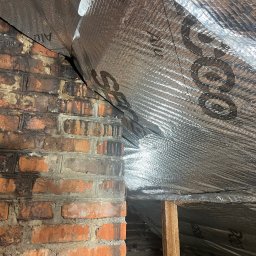 Jakub Stefaniak - Ceglana ściana z widocznymi śladami wilgoci, połączona z izolacją termiczną pokrytą folią aluminiową. Widoczny fragment drewnianej konstrukcji.