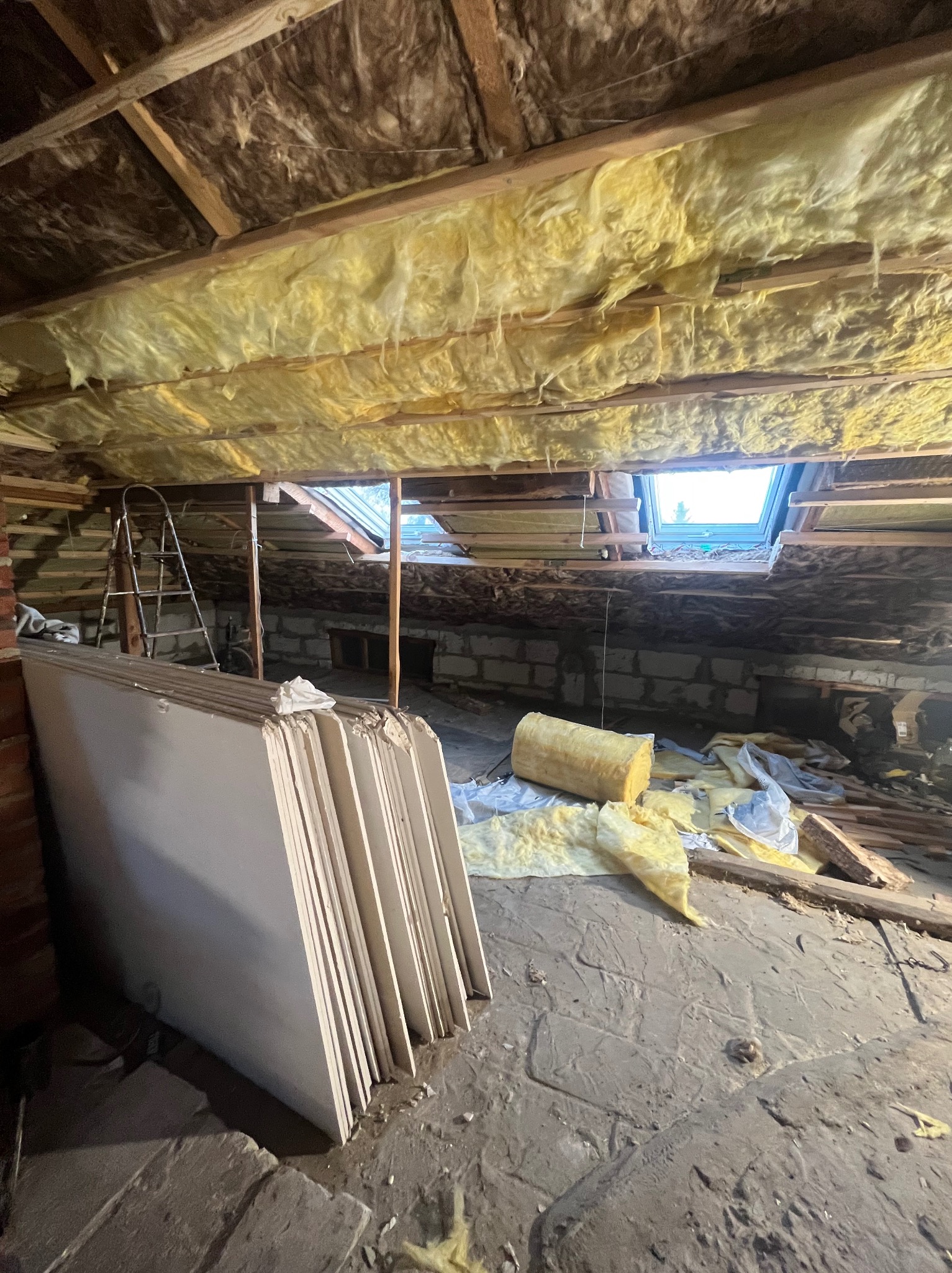 Ocieplone poddasze z oknem dachowym w trakcie prac. Stos płyt kartonowo-gipsowych oparty o ceglaną ścianę. Widoczna drabina i izolacja z wełny mineralnej.