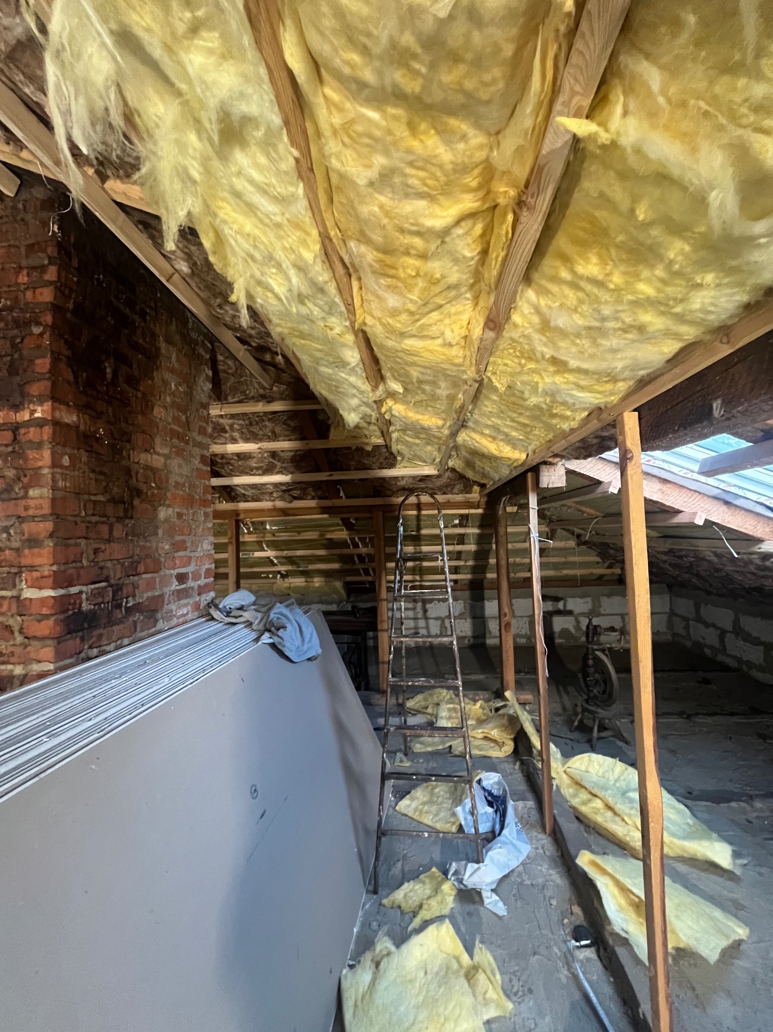 Ocieplenie poddasza wełną mineralną, widoczne płyty gipsowe, drabina i fragment muru z cegły. Prace wykończeniowe na poddaszu.