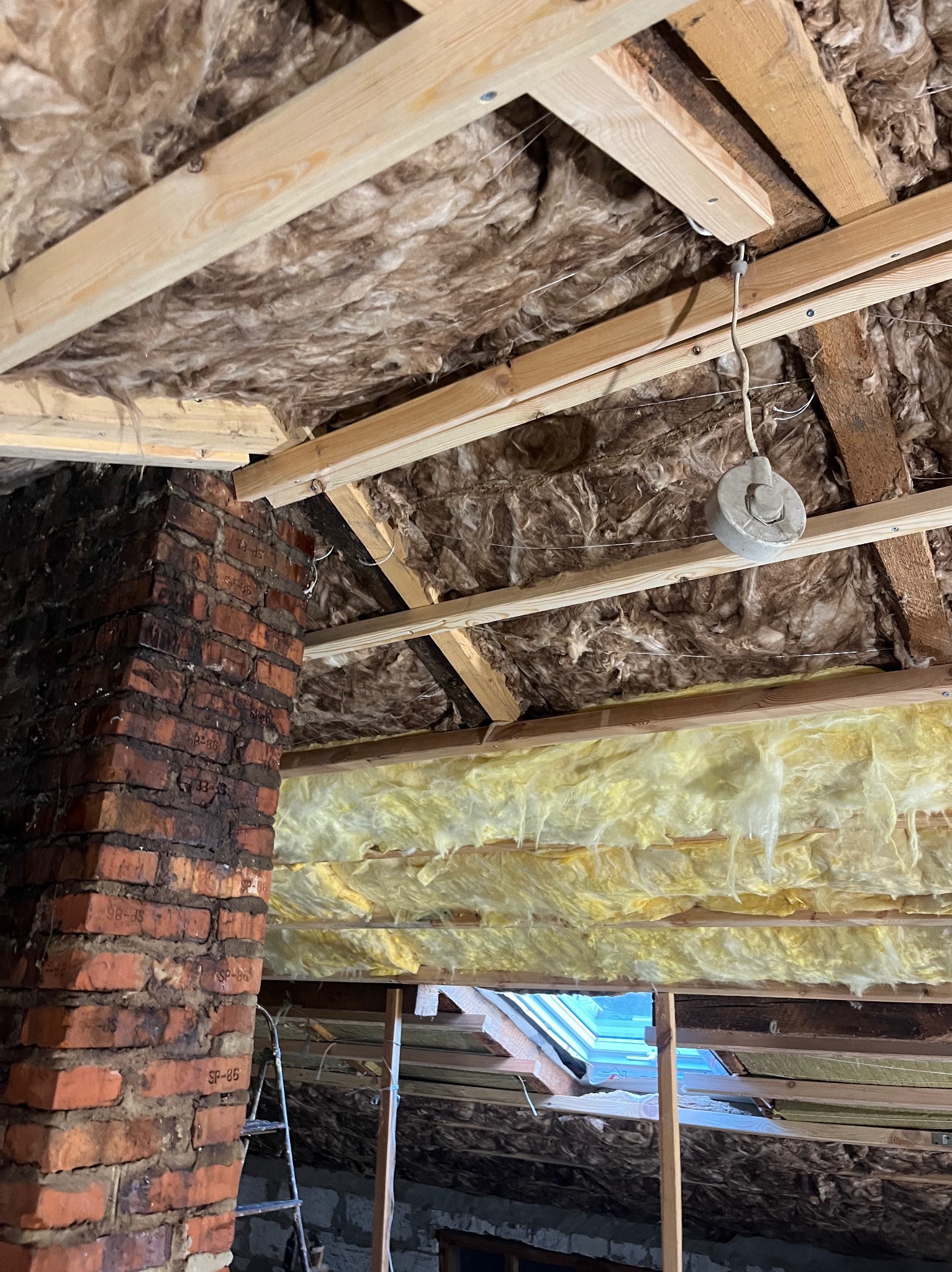 Ocieplenie poddasza wełną mineralną i drewnianą konstrukcją. Widoczny komin z cegły i okno dachowe. Prace wykończeniowe w trakcie realizacji.