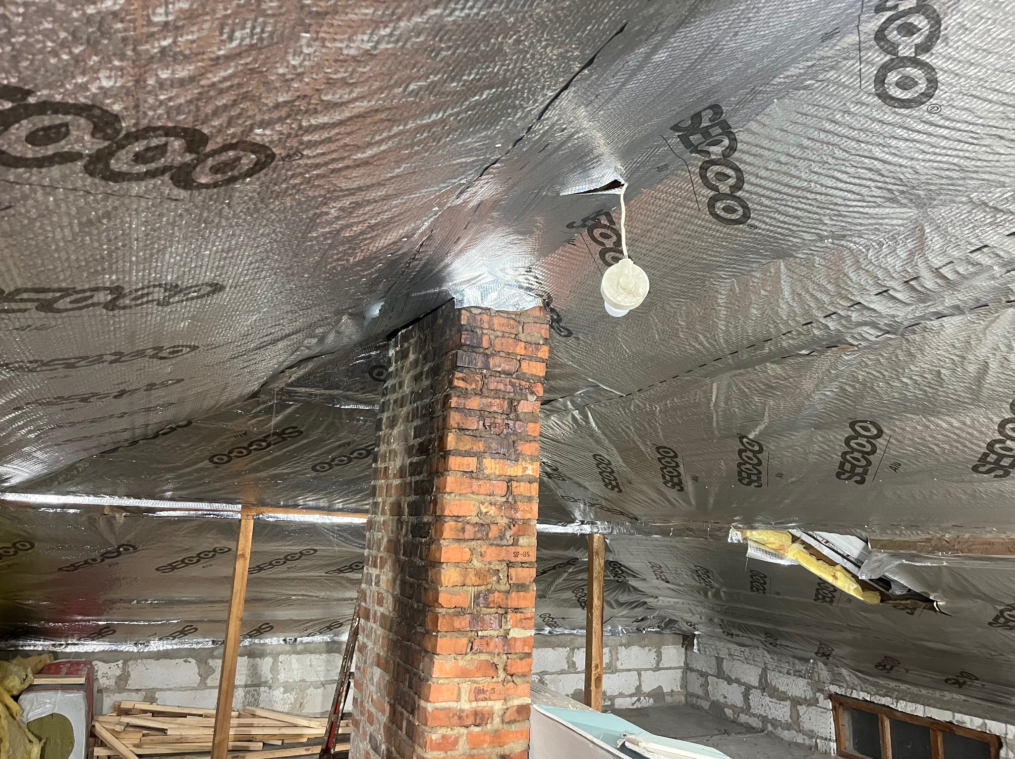 Poddasze w trakcie izolacji folią aluminiową z widocznym kominem z cegły, drewnianymi elementami konstrukcyjnymi i oświetleniem roboczym. Prace remontowe w surowym stanie.