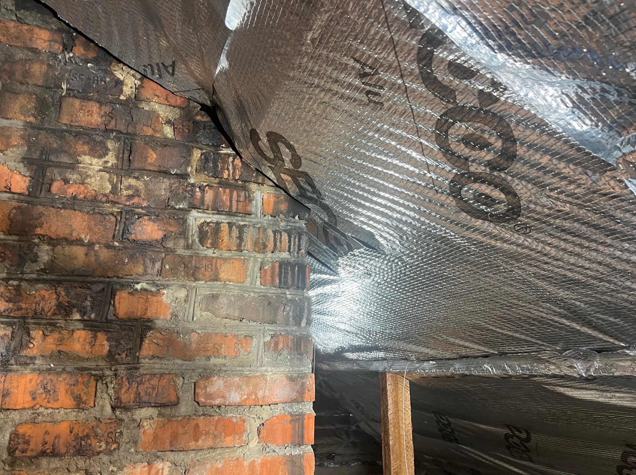 Ceglana ściana z widocznymi śladami wilgoci, połączona z izolacją termiczną pokrytą folią aluminiową. Widoczny fragment drewnianej konstrukcji.